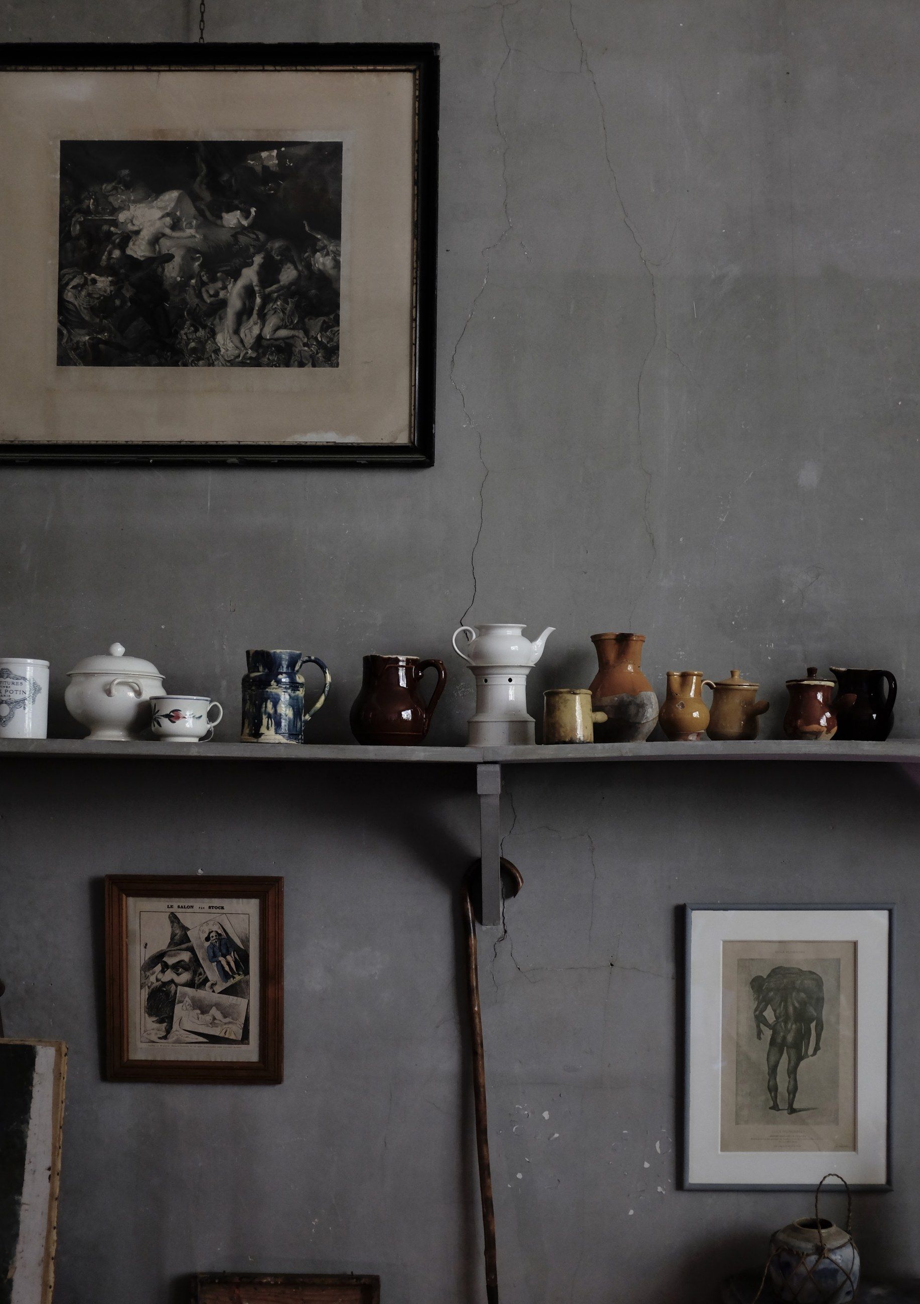 A shelf of jugs and framed prints on a grey wall